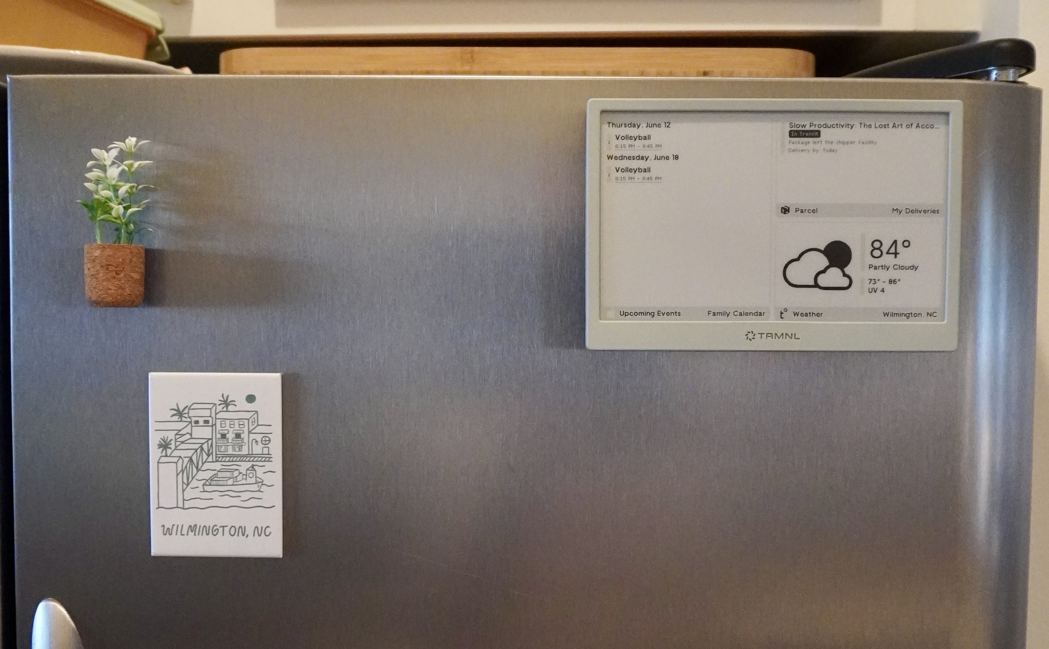 An image of my TRMNL mounted in the top right corner of my fridge. It is display a grid of data, where the left side has our calender and the right side is split between pending deliveries and the current weather. You can also see our Wrightsville Beach, equality, and fake plant magnets in the image